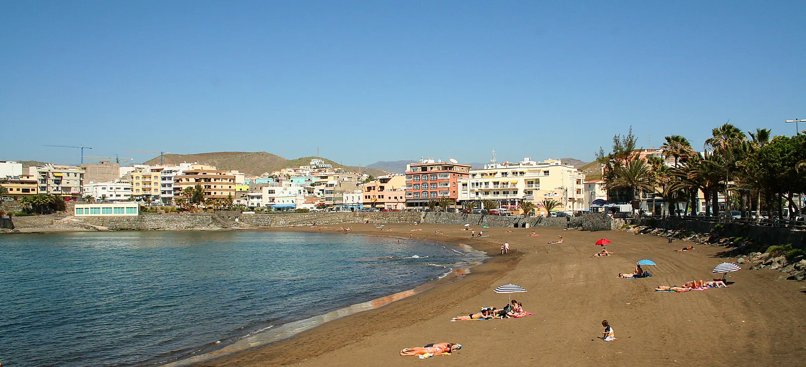 Arguineguín town beach with dark sand, sunbathers and houses climbing the slope under a clear blue sky